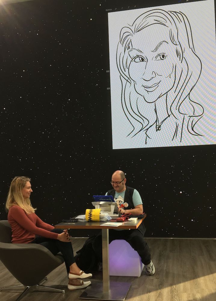 Eine junge Frau sitzt vor Erol auf einer Bühne bei einem SAP-Event in Walldorf, während ein großer Bildschirm hinter ihnen die Entstehung ihrer iPad-Karikatur live zeigt.