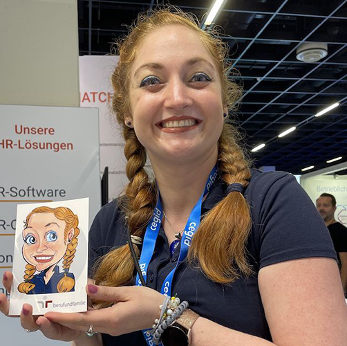Eine junge Frau mit Pippi-Langstrumpf-Zöpfen hält lächelnd ihre iPad-Karikatur auf der Zukunft Personal Europe Messe in Köln in den Händen.