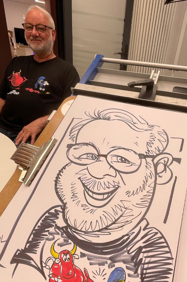 Ein älterer Herr sitzt lächelnd vor Erol bei einer Volksbank-Filialeröffnung nahe Pforzheim; sein lustiges Comic-T-Shirt wird detailgetreu in der iPad-Karikatur übernommen.